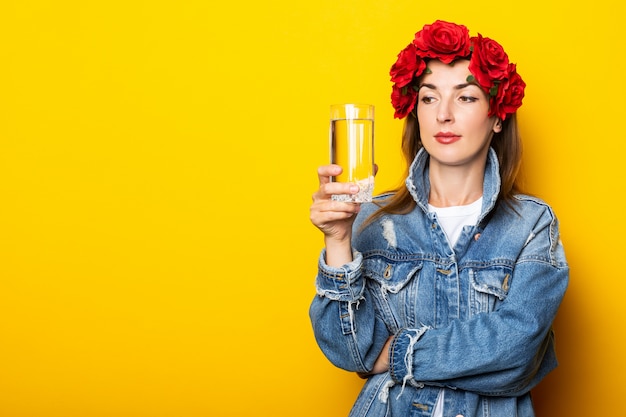 Jonge vrouw in spijkerjasje en een krans van rode bloemen op haar hoofd die een glas water op een gele muur houden.