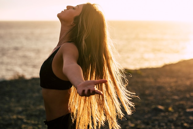 Jonge vrouw in sportkleding met uitgestrekte armen en ogen gesloten staande op het strand, vrouw doet oefening aan de kustlijn. Profiel van mooie zorgeloze sportieve vrouw met lang haar op strand