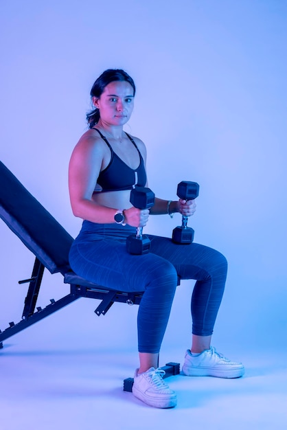 Jonge vrouw in sportkleding zittend op een bankje met twee halters in haar handen