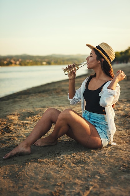 Jonge vrouw in strohoed ontspannen bij zonsondergang op de rivieroever en bier drinken.