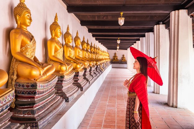 Jonge vrouw in traditionele rode Thaise jurk en gouden accessoires houdt een traditionele paraplu vast in de historische site Wat Phutthaisawan Ayutthaya Thaise nationale kostuum