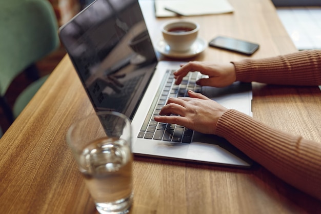 Jonge vrouw in vrijetijdskleding bezig met moderne laptop zittend aan tafel met een kopje koffie en een glas water