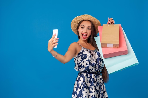 Jonge vrouw in vrijetijdskleding met boodschappentassen die selfiestick gebruiken om een zelfportret te maken op een blauwe studioachtergrond met kopieerruimte