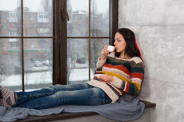 Jonge vrouw in warme gebreide trui liggend op de kussens op de vensterbank en een kopje koffie drinken