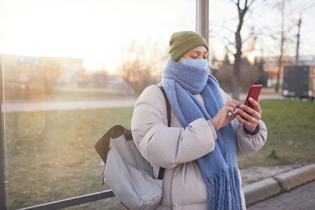 Jonge vrouw in warme kleding en in masker een bericht aan het typen op haar mobiele telefoon tijdens het wachten op de bus