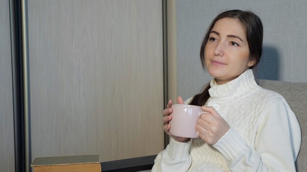 Jonge vrouw in warme trui zittend op de bank en thuis thee drinken.