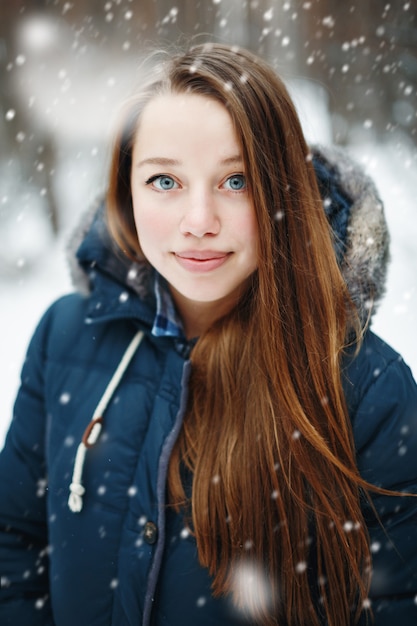 Jonge vrouw in winterkleren staan onder de sneeuw, glimlachen, camera kijken. Winter boslandschap en sneeuwval op de achtergrond.