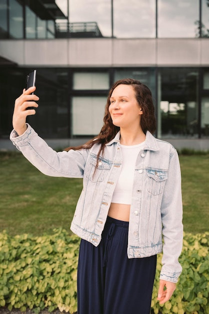 Jonge vrouw in wit spijkerjasje die selfie op straat neemt kantoorgebouw op de achtergrond