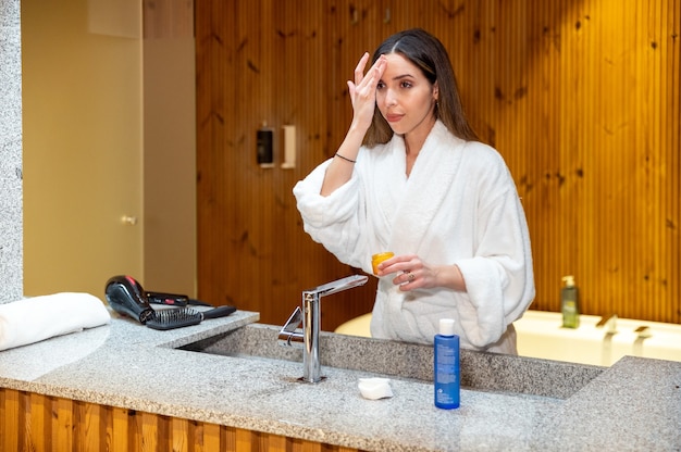 Jonge vrouw in witte badjas staande voor spiegel in de badkamer en cosmetische crème op gezicht toe te passen tijdens de dagelijkse huidverzorgingsroutine