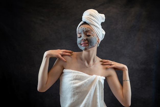 Jonge vrouw in witte handdoek die grijs hydraterend bubbelmasker aanbrengt geïsoleerd op zwart