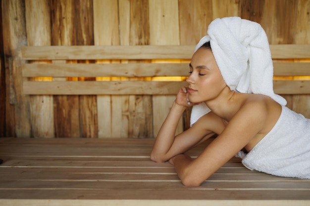 Jonge vrouw in witte handdoek die in houten sauna ligt