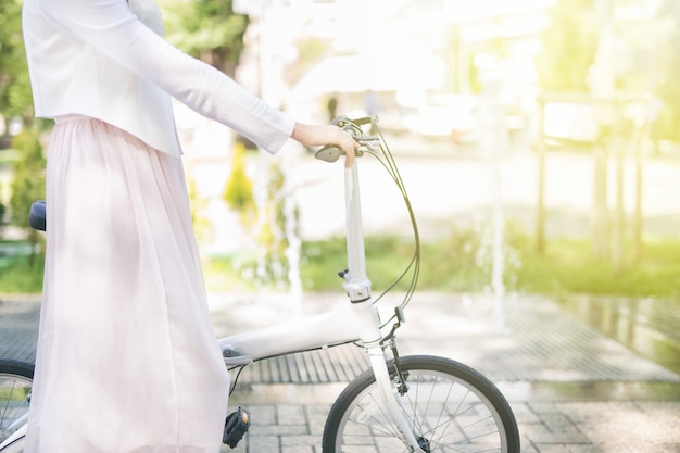 Jonge vrouw in witte jas en roze rok houdt haar hand op het stuur van een witte fiets