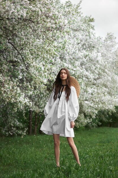 Jonge vrouw in witte jurk met een strohoed loopt door het bloeiende lentetuinpark. De lente is aangebroken, romantische sfeer