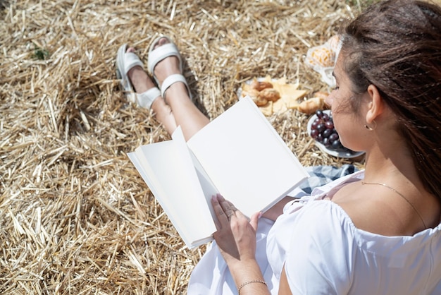 Jonge vrouw in witte jurk zittend op hooiberg in geoogste veld lezen leeg boek boek mockup