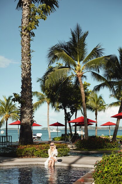 Jonge vrouw in witte peignoir en bikini ontspannen bij het buitenzwembad onder exotische palmbomen ergens in tropisch land. Vakantie en zomer.