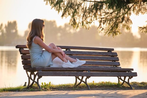 Jonge vrouw in zomerjurk ontspannen zittend op een bankje aan het meer op een warme avond Wellness rustend van het alledaagse haastconcept