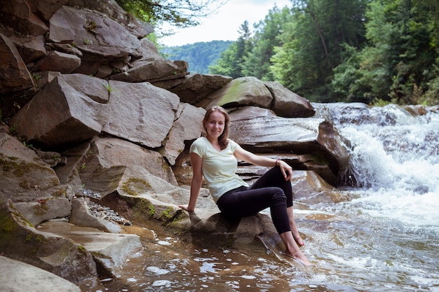 Jonge vrouw in zwarte legging zit op steen in de buurt van splashing waterfall. vreedzame kaukasische reiziger zit bij beautiful stream.