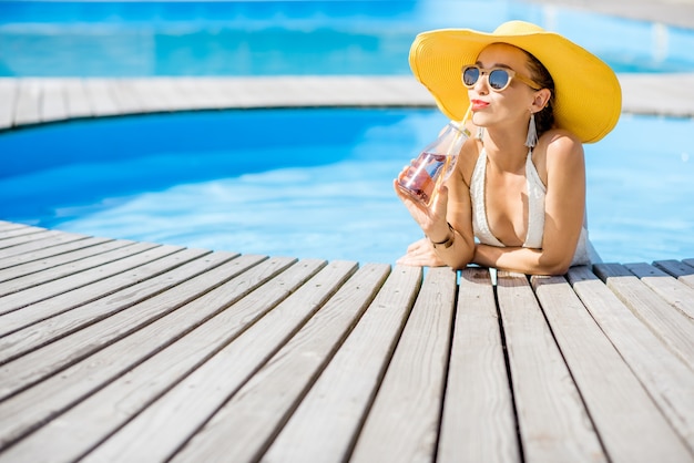 Jonge vrouw in zwembroek met grote gele zonnehoed die ontspant met een fles verse drank die buiten aan het zwembad zit