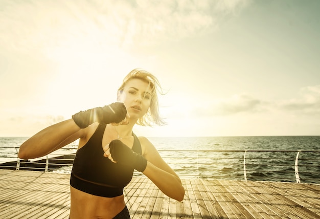 Foto jonge vrouw kickbokser met gewikkelde handen in verband traint een klap op het strand bij zonsopgang. ochtendtraining vechter
