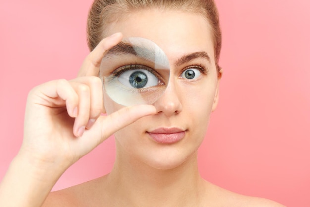 Jonge vrouw kijken naar de camera door vergrootglas met grappige verbaasde uitdrukking, close-up geïsoleerd op roze. Vergroot oog, optische illusie, zoeken