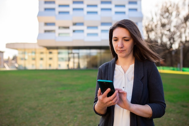 Jonge vrouw kijkt naar de telefoon, praat aan de telefoon, praat op video, glimlacht, zwaait, begroet, op straat in het park, in de buurt van het zakencentrum