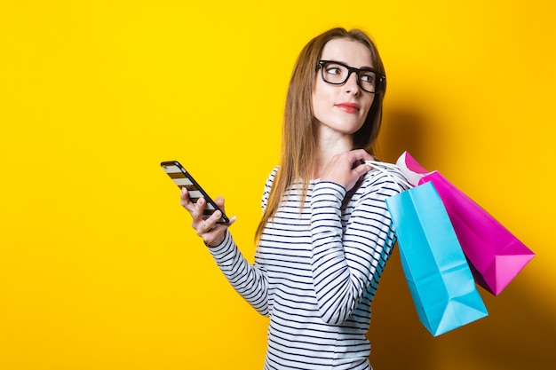 Jonge vrouw kijkt terug, houdt een telefoon en boodschappentassen op een gele achtergrond