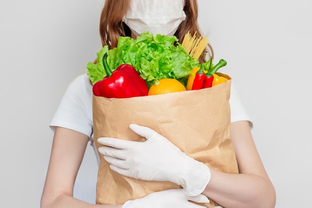 Jonge vrouw koerier vrijwilliger draagt een medisch masker houdt een papieren zak met producten, groenten, chili, kruiden geïsoleerd over wit, grijze ruimte, voedsel levering concept