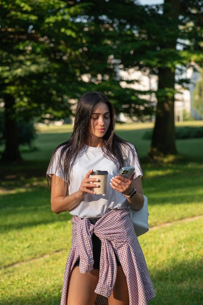 Jonge vrouw koffie drinken glimlachend en kijkend naar de telefoon in het park bij de boom