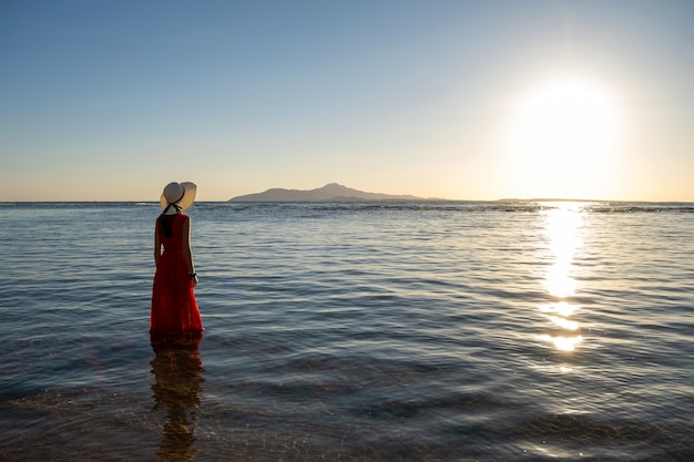 Jonge vrouw lange rode kleding dragen en strohoed die zich in zeewater bevinden bij het strand die van mening van het toenemen zon in de vroege zomerochtend genieten.