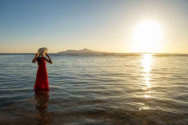 Jonge vrouw lange rode kleding dragen en strohoed die zich in zeewater bevinden bij het strand die van mening van het toenemen zon in de vroege zomerochtend genieten.