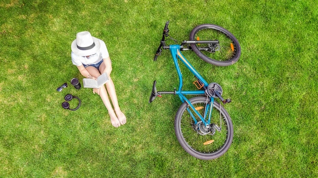 Jonge vrouw leesboek in park in de buurt van fiets. student meisje ontspannen buiten zittend op het gras