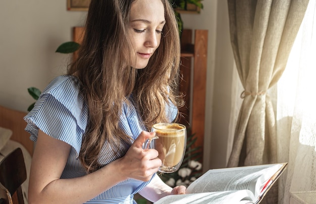 Jonge vrouw leest een boek en drinkt koffie bij het raam Concept van ochtendgenot, vrije tijd aangenaam tijdverdrijf en ontspanning