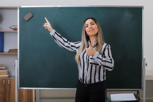 jonge vrouw, leraar, staand, dichtbij, bord, in, klaslokaal, uitleggen, les, wijzend, op, bord, met, wijsvinger, weergeven, duim omhoog, glimlachen, zelfverzekerd