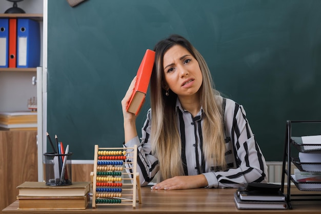 jonge vrouw leraar zit op school bureau voor schoolbord in de klas met een boek dat er moe en overwerkt uitziet