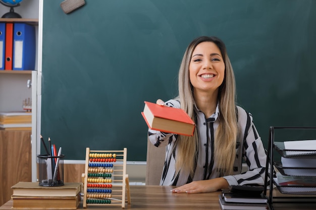 jonge vrouw leraar zit op school bureau voor schoolbord in de klas met een boek dat het aanbiedt aan iemand die vriendelijk lacht