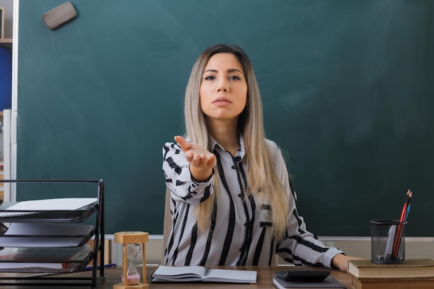 jonge vrouw leraar zit op school bureau voor schoolbord in klas huiswerk controleren van studenten verhogen arm maken kom hier gebaar