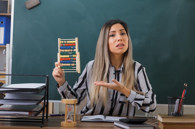 jonge vrouw leraar zit op school bureau voor schoolbord in klaslokaal uitleggen les houden abacus presenteren met arm op zoek verward
