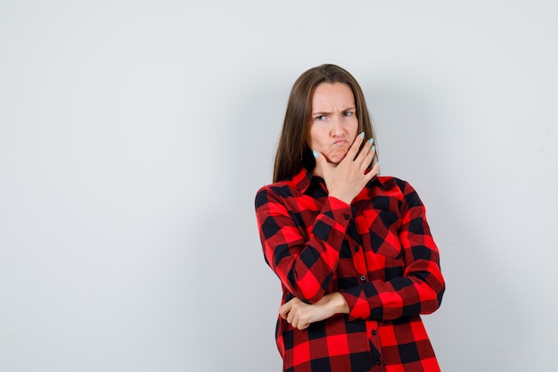 Jonge vrouw leunend met de kin aan de kant, grimassen in geruit overhemd en boos kijken. vooraanzicht.