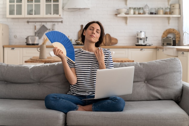 Jonge vrouw lijdt aan zonnesteek plat zonder airconditioner die met ventilator zwaait die thuis op de bank zit