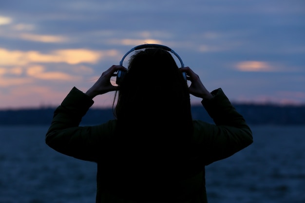 Jonge vrouw luisteren naar muziek in de buurt van de rivier bij zonsondergang