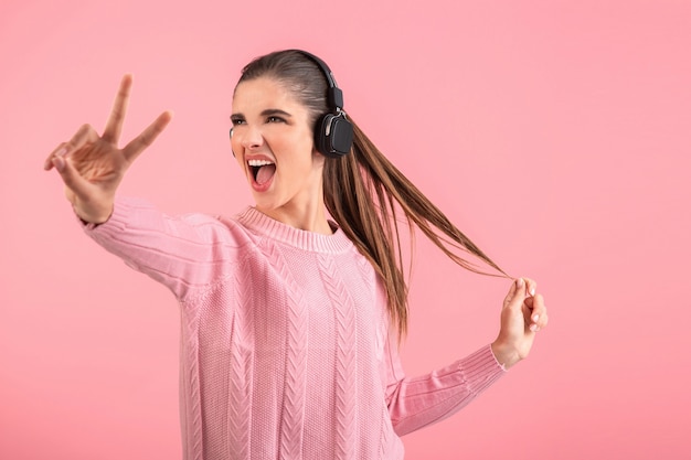 Jonge vrouw luisteren naar muziek in draadloze koptelefoon dragen roze trui glimlachend poseren op roze