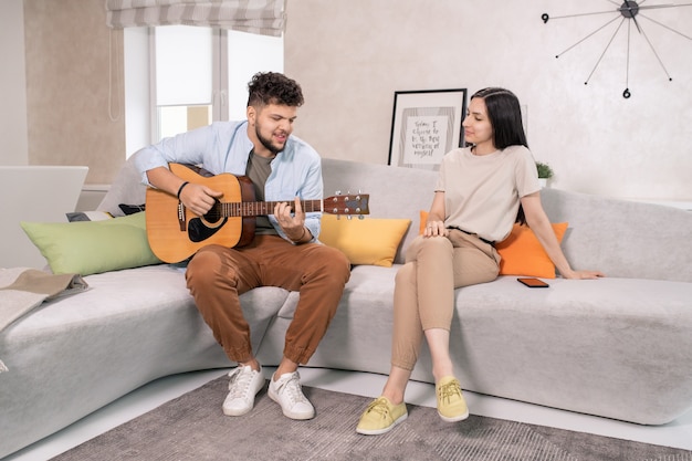 Jonge vrouw luistert naar haar man die gitaar speelt in een thuisomgeving