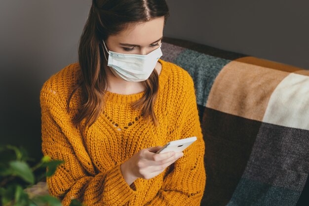 Jonge vrouw maakt gebruik van een mobiele telefoon en zit in een oranje trui in een medisch masker thuis geïsoleerd in quarantaine en leest het nieuws grijze interieur close-up