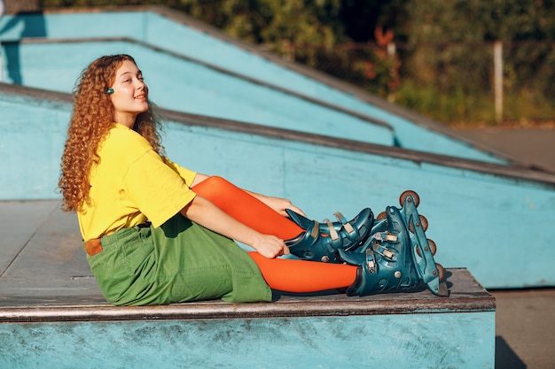 Jonge vrouw meisje in groene en gele kleding en oranje kousen met krullend kapsel rolschaatsen zittend in skatepark