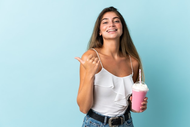 Jonge vrouw met aardbeimilkshake die op blauw wordt geïsoleerd dat naar de kant wijst om een product te presenteren