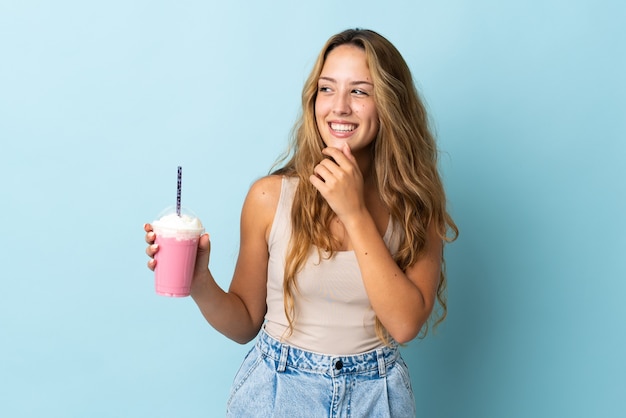 Jonge vrouw met aardbeimilkshake die op blauwe muur wordt geïsoleerd die aan de kant kijkt en glimlacht