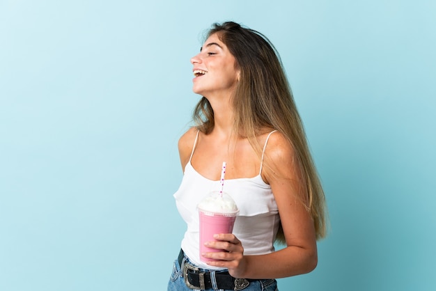 Jonge vrouw met aardbeimilkshake die op blauwe muur wordt geïsoleerd die in zijpositie lacht