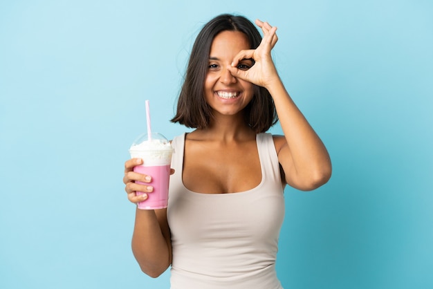 Jonge vrouw met aardbeimilkshake die op blauwe muur wordt geïsoleerd die ok teken met vingers toont