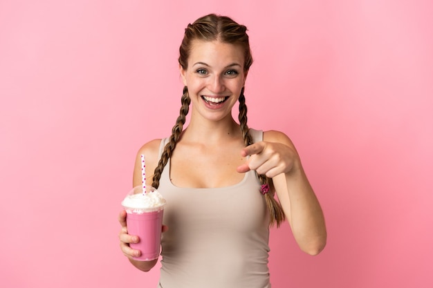 Jonge vrouw met aardbeimilkshake geïsoleerd op roze verrast en wijzend front