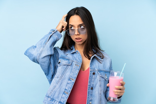 Jonge vrouw met aardbeimilkshake op geïsoleerd blauw die twijfels hebben en denken